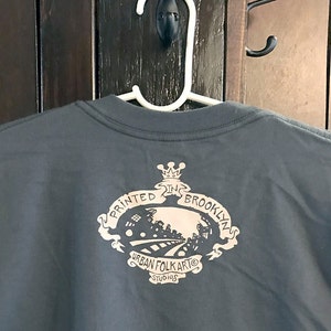 May include: A dark gray t-shirt with a black and white graphic print on the back. The graphic features a crown, the text "Printed in Brooklyn", and the text "Urban Folk Art Studios".