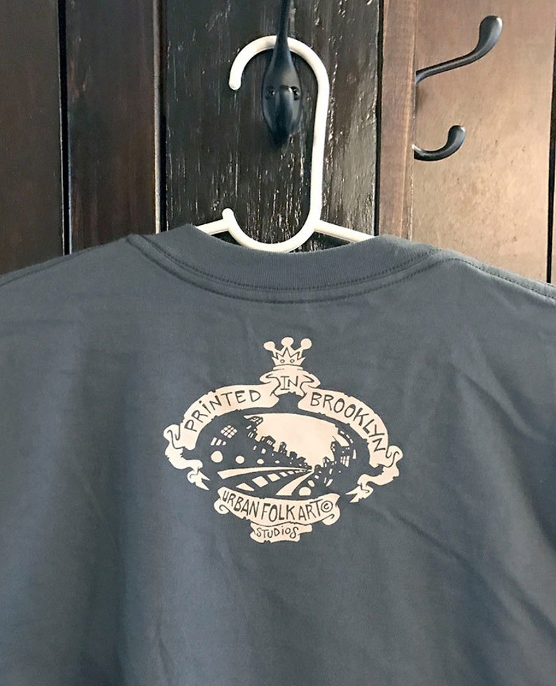 May include: A dark gray t-shirt with a black and white graphic print on the back. The graphic features a crown, the text "Printed in Brooklyn", and the text "Urban Folk Art Studios".