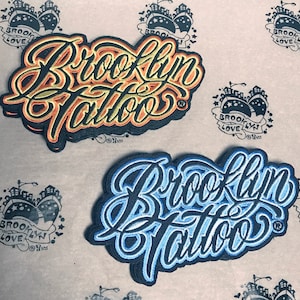 May include: Two embroidered patches with the text "Brooklyn Tattoo" in a stylized font. The top patch is orange and yellow, while the bottom patch is blue and white.