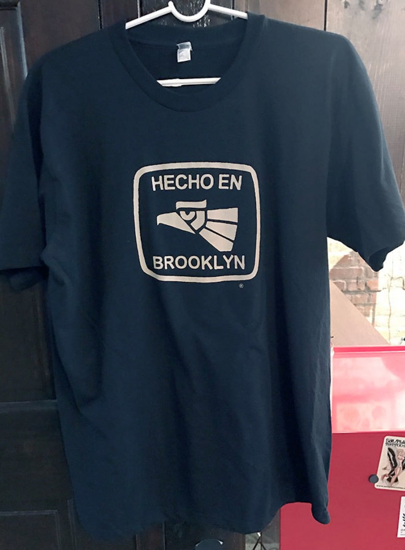 May include: Black t-shirt with a white graphic of a stylized eagle and the text "Hecho en Brooklyn" in a white square.