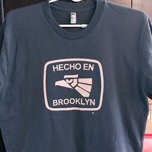 May include: A dark blue t-shirt with a white graphic of a stylized eagle head inside a square with the text "HECHO EN BROOKLYN" in white.