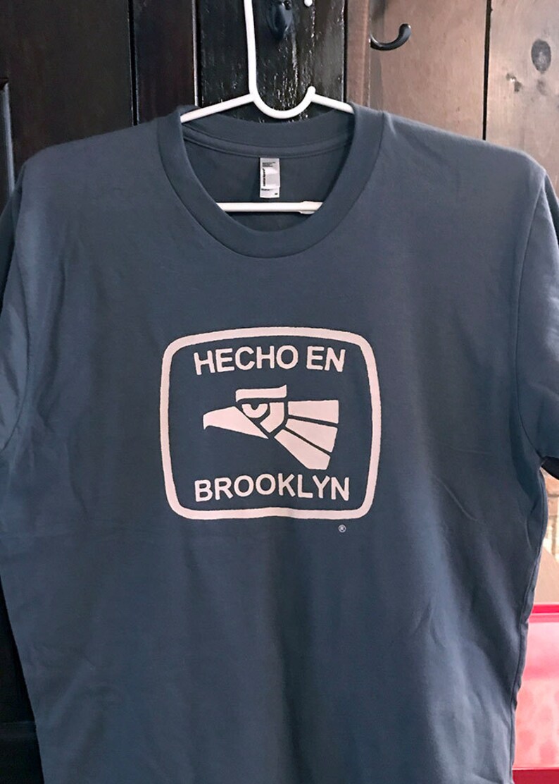 May include: A dark blue t-shirt with a white graphic of a stylized eagle head inside a square with the text "HECHO EN BROOKLYN" in white.