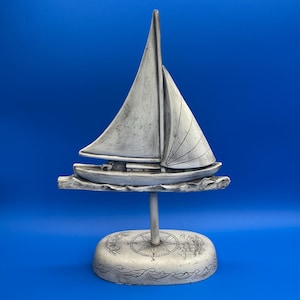 Puede incluir: Una detallada escultura de velero gris con un mástil alto y velas ondeantes. El barco se asienta sobre una base texturizada con un diseño de rosa de los vientos y detalles de olas. La escultura está sostenida por una fina varilla metálica, sobre un fondo azul vibrante.