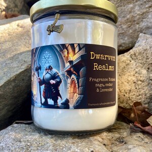 May include: A glass jar candle with a gold lid and a small axe charm. The candle is white and has a label that says "Dwarven Realms" and "Fragrance Notes: sage, cedar & lavender". The label also includes the text "TheHandcraftedHobbit.com".