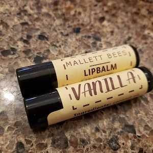 May include: Two brown and black lip balm tubes with a yellow label. The label says "Mallett Bees Lipbalm Vanilla" and "Ingredients: Coconut oil".