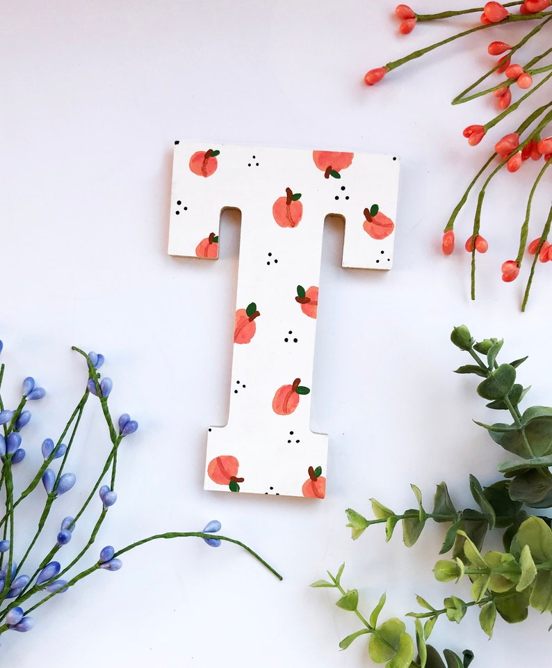 May include: Wooden letter "T" painted white with a pattern of peach-colored fruit and small black dots. The letter is surrounded by sprigs of greenery and flowers against a white background.
