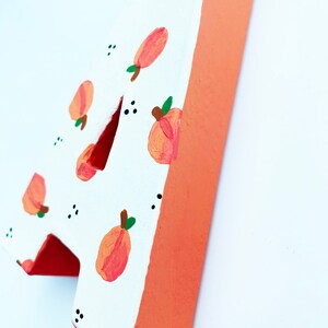 May include: Wooden number four, painted white with a pattern of peaches and green leaves. The edges are painted a solid orange color. Small black dots are scattered around the peach pattern.