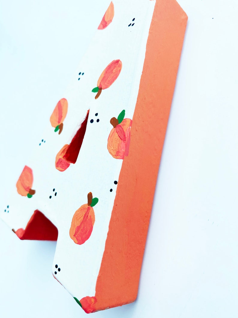 May include: Wooden number four, painted white with a pattern of peaches and green leaves. The edges are painted a solid orange color. Small black dots are scattered around the peach pattern.