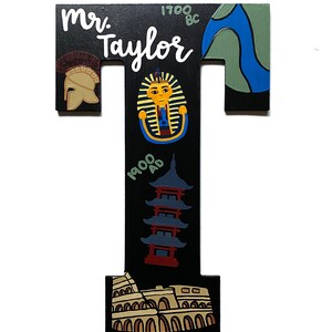 May include: A wooden letter "T" with a black background, featuring illustrations and text. The letter includes a Roman helmet, a pharaoh, a pagoda, and the Colosseum. The name "Mr. Taylor" is written in white.