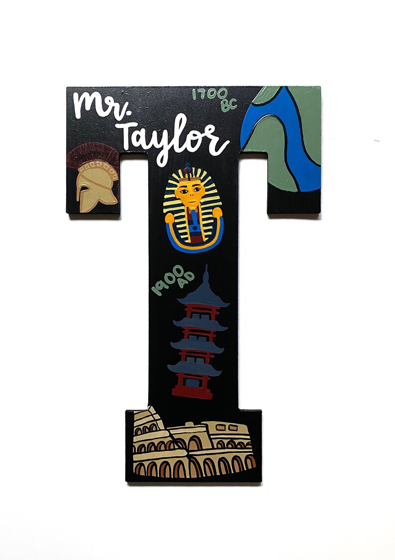May include: A wooden letter "T" with a black background, featuring illustrations and text. The letter includes a Roman helmet, a pharaoh, a pagoda, and the Colosseum. The name "Mr. Taylor" is written in white.
