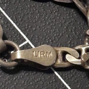 Puede incluir: Una pulsera de cadena de metal plateado con eslabones ovalados y un cierre de mosquet&oacute;n. El cierre tiene grabadas las letras LIRM. La superficie met&aacute;lica muestra un acabado mate ligeramente deslustrado con signos visibles de desgaste.