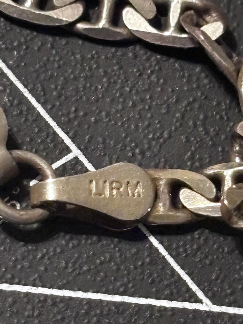 Puede incluir: Una pulsera de cadena de metal plateado con eslabones ovalados y un cierre de mosquet&oacute;n. El cierre tiene grabadas las letras LIRM. La superficie met&aacute;lica muestra un acabado mate ligeramente deslustrado con signos visibles de desgaste.