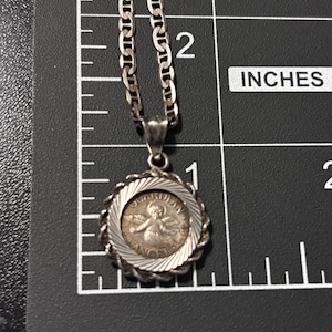 Puede incluir: Un collar con colgante plateado que presenta un dije circular con una figura de &aacute;ngel en relieve y el texto "GUARDIAN ANGEL" en el borde, colocado sobre una regla que muestra un di&aacute;metro aproximado de 1,9 cm.