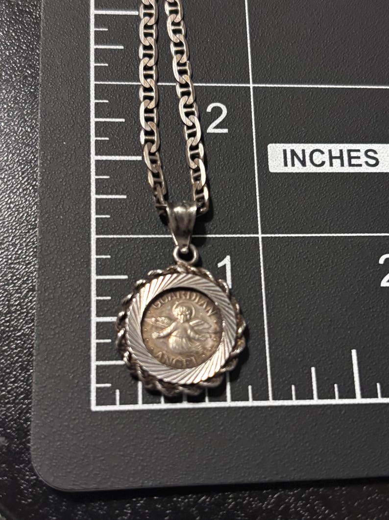 Puede incluir: Un collar con colgante plateado que presenta un dije circular con una figura de &aacute;ngel en relieve y el texto "GUARDIAN ANGEL" en el borde, colocado sobre una regla que muestra un di&aacute;metro aproximado de 1,9 cm.