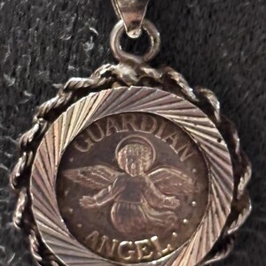 Puede incluir: Un colgante circular de tono plateado que presenta una figura central en relieve de un &aacute;ngel con alas y halo. El texto "GUARDIAN ANGEL" est&aacute; inscrito alrededor de la figura. El colgante tiene un borde de estilo cuerda trenzada y una argolla.