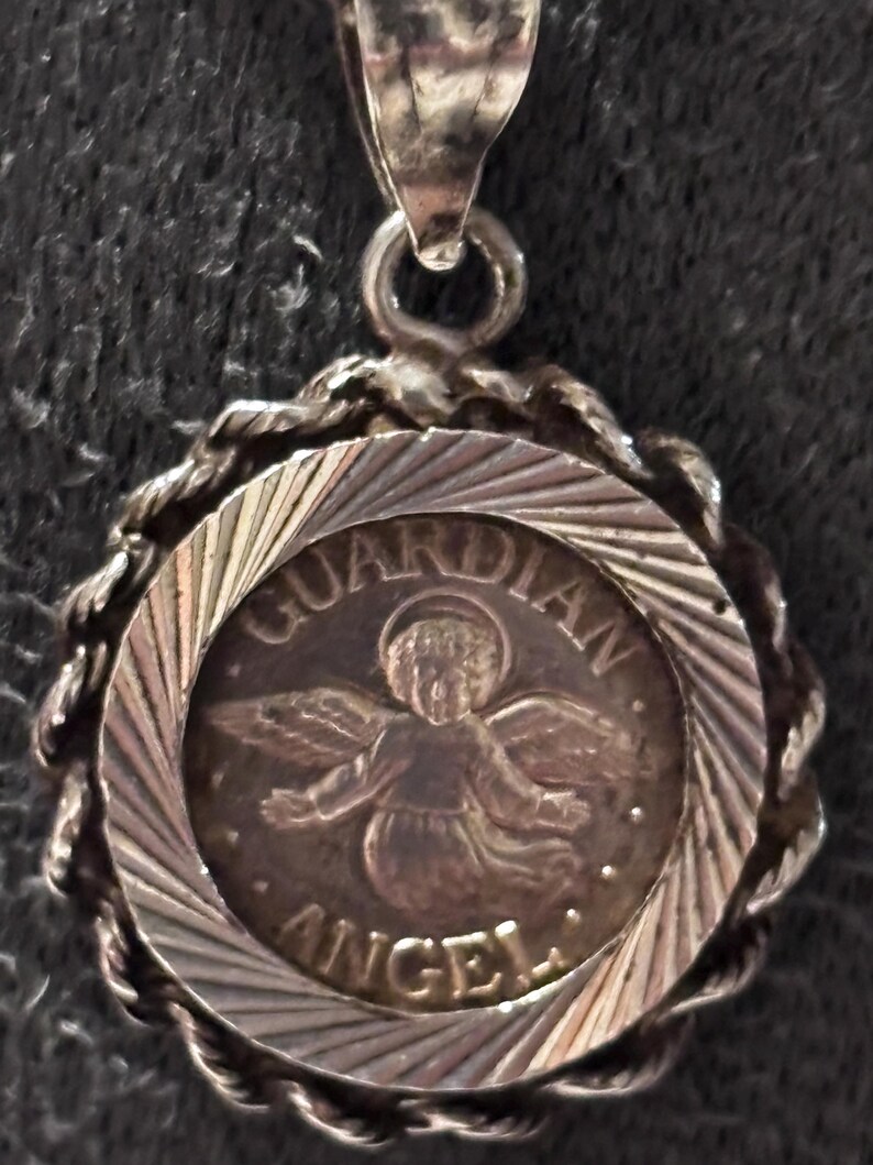 Puede incluir: Un colgante circular de tono plateado que presenta una figura central en relieve de un &aacute;ngel con alas y halo. El texto "GUARDIAN ANGEL" est&aacute; inscrito alrededor de la figura. El colgante tiene un borde de estilo cuerda trenzada y una argolla.