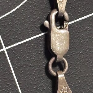 Puede incluir: Un collar de cadena de plata con un cierre de mosquet&oacute;n. Los componentes met&aacute;licos est&aacute;n marcados con 925, ITALY y LIRM. Los eslabones de la cadena son visibles y el mecanismo del cierre se muestra en detalle sobre un fondo oscuro texturizado.