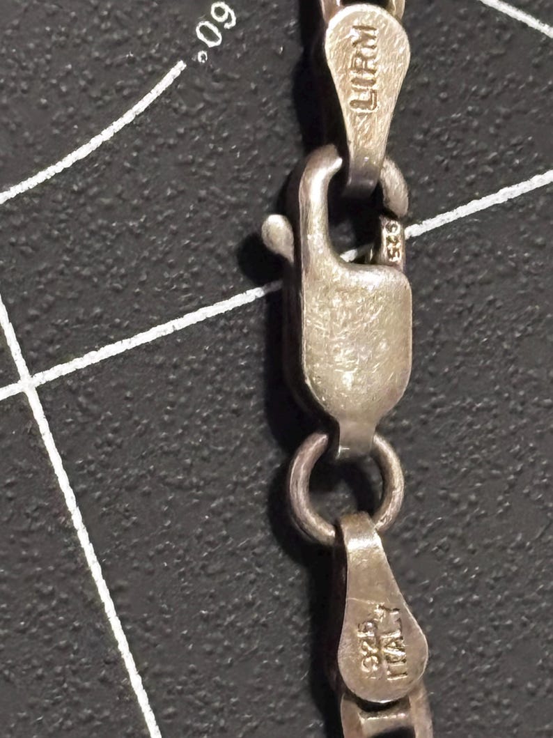 Puede incluir: Un collar de cadena de plata con un cierre de mosquet&oacute;n. Los componentes met&aacute;licos est&aacute;n marcados con 925, ITALY y LIRM. Los eslabones de la cadena son visibles y el mecanismo del cierre se muestra en detalle sobre un fondo oscuro texturizado.