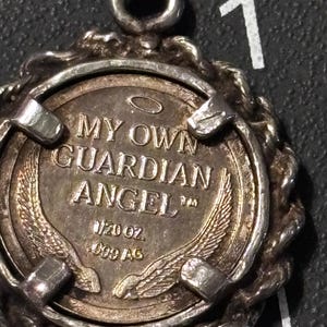 Puede incluir: Un colgante circular de tono plateado con una moneda central que presenta el texto MY OWN GUARDIAN ANGEL, una aureola y dos alas. La moneda est&aacute; marcada con 1/20 OZ y 999 AG, sujeta por un marco de metal trenzado con una anilla en la parte superior.