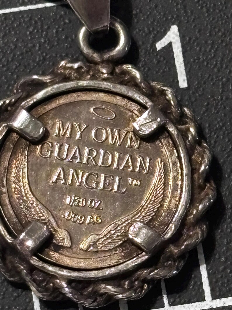 Puede incluir: Un colgante circular de tono plateado con una moneda central que presenta el texto MY OWN GUARDIAN ANGEL, una aureola y dos alas. La moneda est&aacute; marcada con 1/20 OZ y 999 AG, sujeta por un marco de metal trenzado con una anilla en la parte superior.