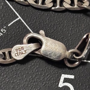 Puede incluir: Un collar de cadena de color plateado con un cierre de mosquet&oacute;n. El cierre est&aacute; estampado con el texto 925 e ITALY. La cadena consta de eslabones ovalados conectados por una barra central, creando un dise&ntilde;o de estilo marinero con acabado met&aacute;lico.