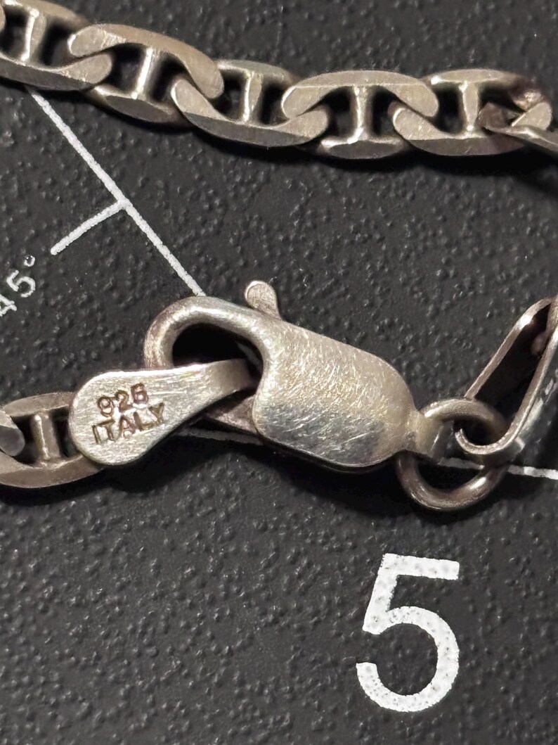Puede incluir: Un collar de cadena de color plateado con un cierre de mosquet&oacute;n. El cierre est&aacute; estampado con el texto 925 e ITALY. La cadena consta de eslabones ovalados conectados por una barra central, creando un dise&ntilde;o de estilo marinero con acabado met&aacute;lico.