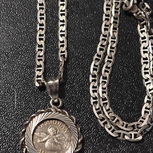 Puede incluir: Un collar de tono plateado con un colgante redondo y un borde estilo cuerda. El centro del colgante muestra una figura alada con el texto "GUARDIAN ANGEL" grabado alrededor del borde. La cadena consta de eslabones ovalados planos entrelazados.