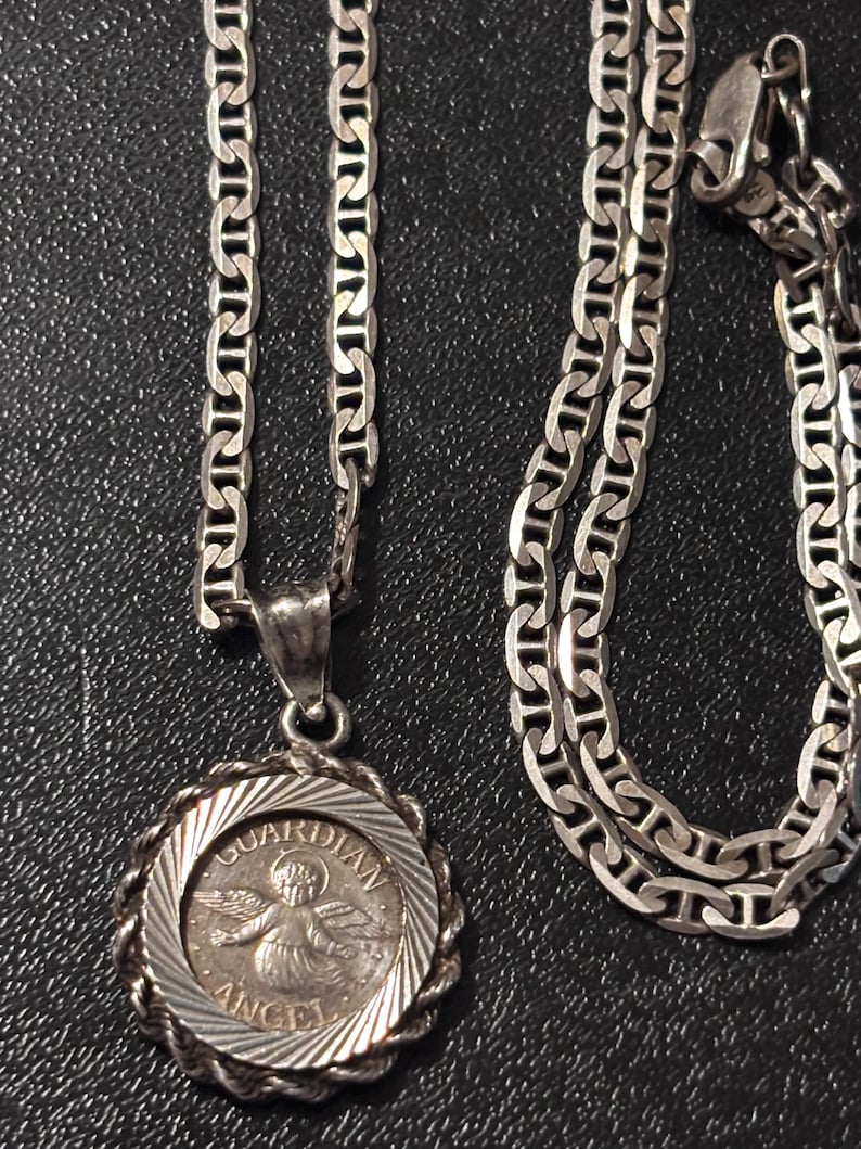 Puede incluir: Un collar de tono plateado con un colgante redondo y un borde estilo cuerda. El centro del colgante muestra una figura alada con el texto "GUARDIAN ANGEL" grabado alrededor del borde. La cadena consta de eslabones ovalados planos entrelazados.