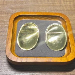 May include: Two gold-colored, oval-shaped earrings are displayed on a dark gray surface within a wooden tray. The earrings have a reflective, polished finish. The tray has a square shape with rounded corners and a warm, brown wood grain.