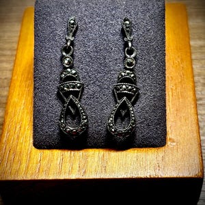 May include: A pair of antique silver-toned drop earrings with a marcasite design. The earrings are displayed on a gray velvet surface inside a wooden box. The earrings have a teardrop shape and are intricately detailed.
