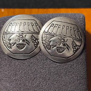 May include: Two silver-toned, round earrings with an embossed design. Each earring features a detailed depiction of a bee, wheat, and a geometric pattern. The earrings are displayed on a dark gray surface.