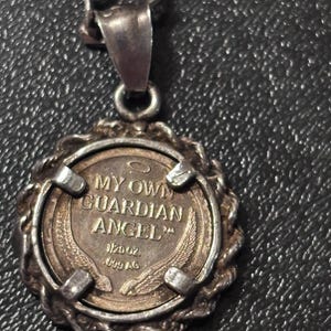 Puede incluir: Un colgante plateado en una cadena con un inserto circular similar a una moneda con el texto MY OWN GUARDIAN ANGEL, un peque&ntilde;o icono de halo y dos alas. La moneda est&aacute; marcada con 1/20 oz .999 AG y se sujeta mediante cuatro garras met&aacute;licas.