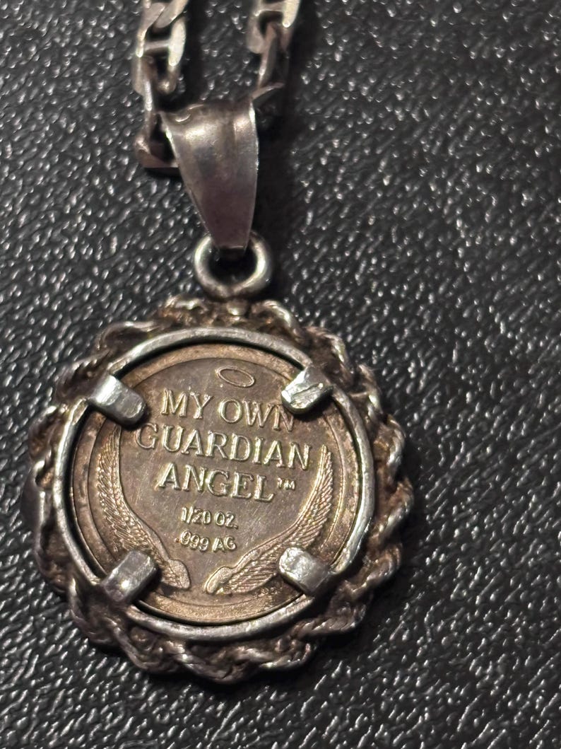 Puede incluir: Un colgante plateado en una cadena con un inserto circular similar a una moneda con el texto MY OWN GUARDIAN ANGEL, un peque&ntilde;o icono de halo y dos alas. La moneda est&aacute; marcada con 1/20 oz .999 AG y se sujeta mediante cuatro garras met&aacute;licas.