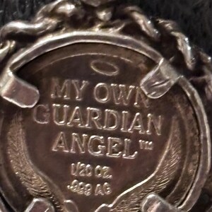 Puede incluir: Un colgante circular de tono plateado con un borde estilo cuerda y un disco central. El disco est&aacute; grabado con el texto MY OWN GUARDIAN ANGEL, un s&iacute;mbolo de halo, las inscripciones 1/20 oz. y .999 AG, junto con im&aacute;genes de alas estilizadas.