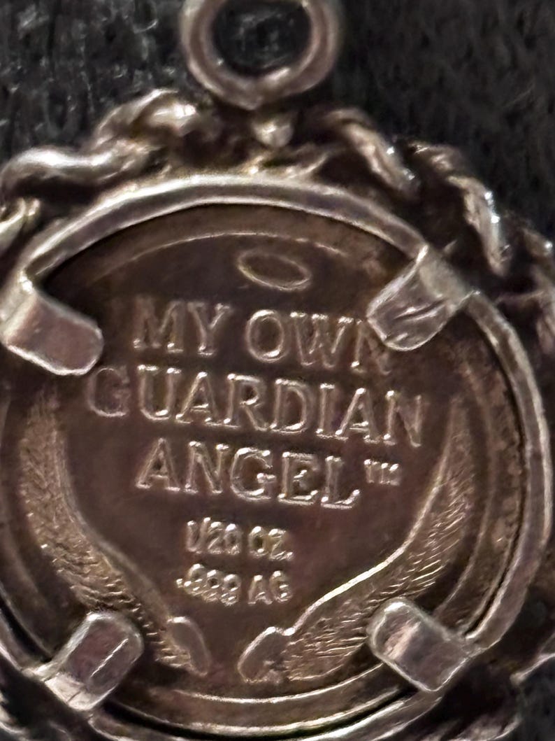 Puede incluir: Un colgante circular de tono plateado con un borde estilo cuerda y un disco central. El disco est&aacute; grabado con el texto MY OWN GUARDIAN ANGEL, un s&iacute;mbolo de halo, las inscripciones 1/20 oz. y .999 AG, junto con im&aacute;genes de alas estilizadas.