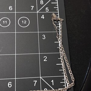 Puede incluir: Un collar de cadena de metal plateado con eslabones planos de forma ovalada, colocado sobre una alfombrilla de medici&oacute;n negra con l&iacute;neas de cuadr&iacute;cula y n&uacute;meros blancos. La cadena se extiende desde la esquina superior derecha hacia abajo, midiendo m&aacute;s de 15 cm.