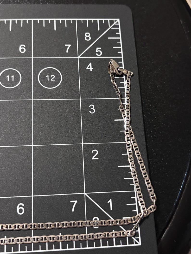 Puede incluir: Un collar de cadena de metal plateado con eslabones planos de forma ovalada, colocado sobre una alfombrilla de medici&oacute;n negra con l&iacute;neas de cuadr&iacute;cula y n&uacute;meros blancos. La cadena se extiende desde la esquina superior derecha hacia abajo, midiendo m&aacute;s de 15 cm.