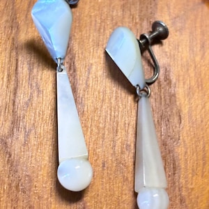 May include: A pair of vintage earrings with a silver-tone metal setting. Each earring features a light blue, faceted top, a white, elongated, triangular middle section, and a white, spherical bottom. The earrings are displayed on a wooden surface.