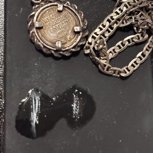 Puede incluir: Un collar con colgante de color plateado que presenta un dije circular estilo moneda con un borde de textura de cuerda. El dije tiene grabada la inscripci&oacute;n MY OWN GUARDIAN ANGEL, un peque&ntilde;o icono de halo y tallos de trigo debajo.