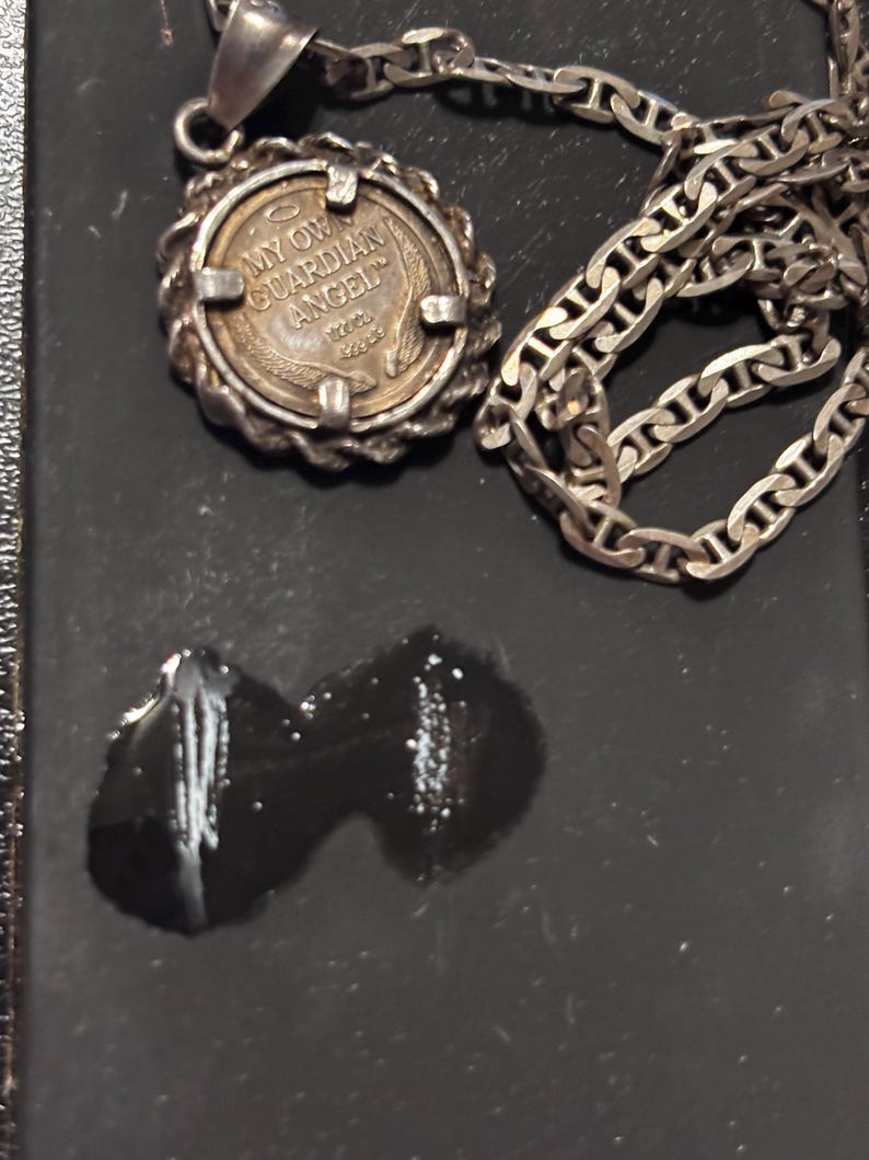 Puede incluir: Un collar con colgante de color plateado que presenta un dije circular estilo moneda con un borde de textura de cuerda. El dije tiene grabada la inscripci&oacute;n MY OWN GUARDIAN ANGEL, un peque&ntilde;o icono de halo y tallos de trigo debajo.