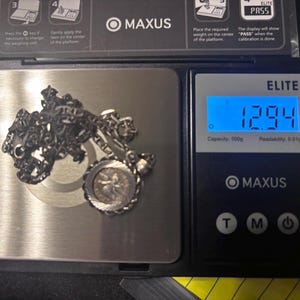 Puede incluir: Un collar de cadena de tono plateado con un colgante circular que presenta una figura central, descansando sobre una b&aacute;scula digital que muestra un peso de 12,94 gramos. La b&aacute;scula es un modelo Maxus Elite negro con plataforma met&aacute;lica.