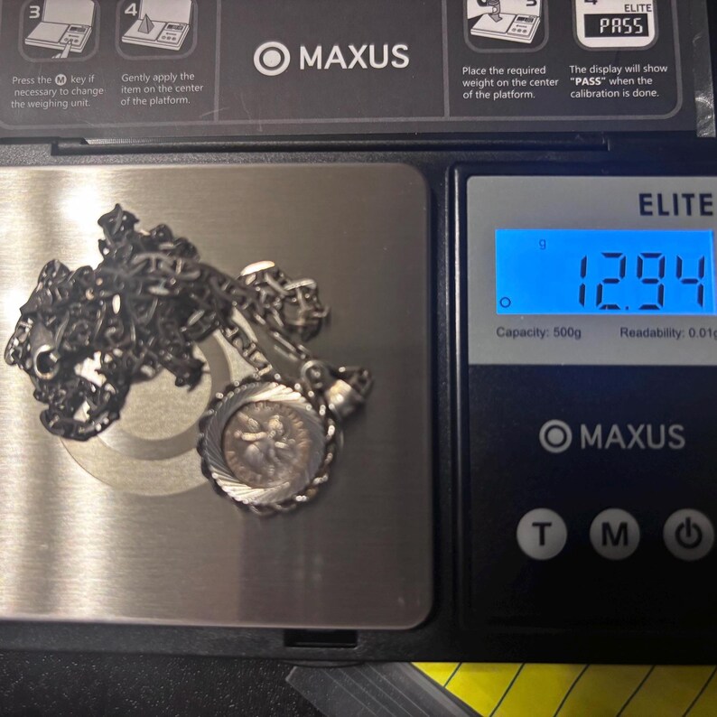 Puede incluir: Un collar de cadena de tono plateado con un colgante circular que presenta una figura central, descansando sobre una b&aacute;scula digital que muestra un peso de 12,94 gramos. La b&aacute;scula es un modelo Maxus Elite negro con plataforma met&aacute;lica.