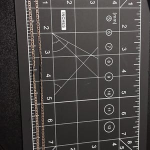 Puede incluir: Un collar de cadena de metal de color plateado con un dise&ntilde;o de eslabones planos, colocado verticalmente sobre una alfombrilla de medici&oacute;n negra. La cadena se extiende desde cero hasta unos 19 cm de largo y tiene un peque&ntilde;o cierre en el extremo.