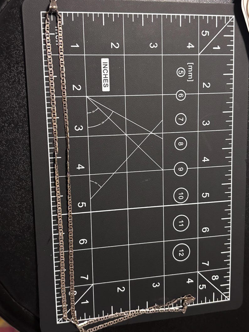 Puede incluir: Un collar de cadena de metal de color plateado con un dise&ntilde;o de eslabones planos, colocado verticalmente sobre una alfombrilla de medici&oacute;n negra. La cadena se extiende desde cero hasta unos 19 cm de largo y tiene un peque&ntilde;o cierre en el extremo.