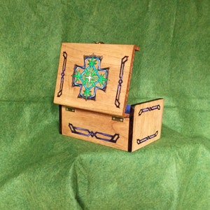 May include: A wooden box with a hinged lid, decorated with a blue and green stained glass design of a cross. The box is closed and sitting on a green surface.