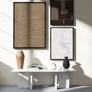 May include: Three abstract art prints in black frames hang on a white wall above a white marble table with a brown vase, a white ceramic vase, and a black vase. The table is in a room with a light grey floor and a window letting in natural light.