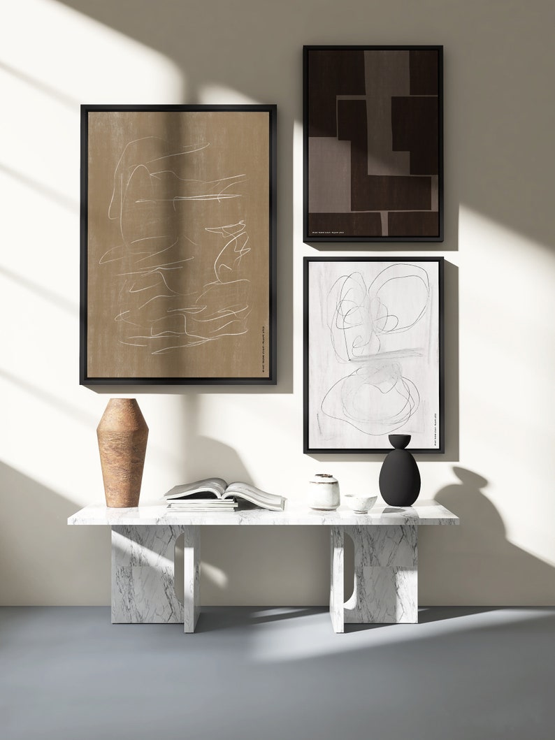 May include: Three abstract art prints in black frames hang on a white wall above a white marble table with a brown vase, a white ceramic vase, and a black vase. The table is in a room with a light grey floor and a window letting in natural light.