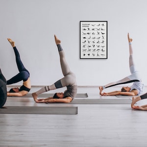 May include: Three people are practicing pilates on gray yoga mats in a white room. A framed poster with the text "Pilates Mat Exercises" and a series of black silhouettes of pilates poses is hanging on the wall.