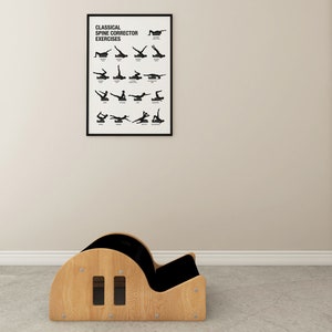 May include: A wooden Pilates spine corrector with a black cushioned seat and a framed poster displaying classical spine corrector exercises. The poster is black and white and hangs on a beige wall.