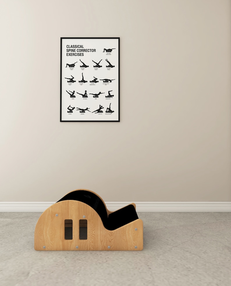 May include: A wooden Pilates spine corrector with a black cushioned seat and a framed poster displaying classical spine corrector exercises. The poster is black and white and hangs on a beige wall.